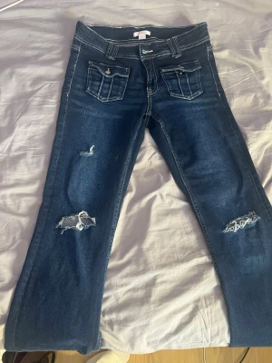Mörkblå slitna bootcut jeans - Säljer ett par mörkblå bootcut jeans med slitningar och råa detaljer på benen. Jeansen har dubbla framfickor med lock och kontrastsömmar som ger en snygg look. Perfekta för dig som gillar en avslappnad och trendig stil.