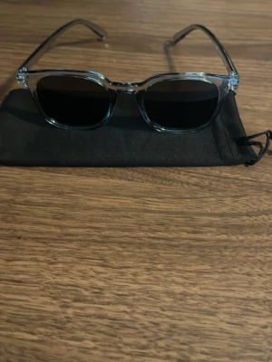 Chimi liknande solglasögon - Säljer ett par stilrena solglasögon med transparent båge och svarta glas. Bågarna har en modern fyrkantig form och kommer med en svart tygpåse. Perfekta för att lyfta din look under soliga dagar. Ska ha uv skydd men har inte testar för uv skydd