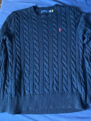 Mörkblå kabelstickad tröja Polo Ralph Lauren - Kabelstickad mörkblå tröja från Polo Ralph Lauren med rund halsringning och ribbade muddar. Klassisk röd logga broderad på bröstet. Tröjan har lång ärm och är tillverkad i mjukt material som passar perfekt till höst och vinter.