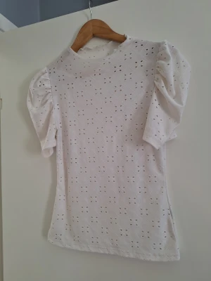 Vit blus med puffärm och broderi - Använd fåtal gånger. Superfin vit blus med broderade hålmönster och puffiga ärmar. Toppen har rund halsringning och är tillverkad i ett lätt bomullstyg som känns luftigt. Perfekt för dig som gillar romantiska detaljer och en feminin stil.