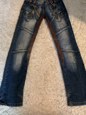Unika blå jeans med bruna detaljer - Säljer ett par blå jeans med slitningar och bruna tygpaneler längs insidan av benen och grenen. Jeansen har flera dragkedjor och dekorativa sömmar framtill samt fickor både fram och bak. Materialet är denim med inslag av brunt tyg för en cool kontrast. Ger en cowboy känsla  