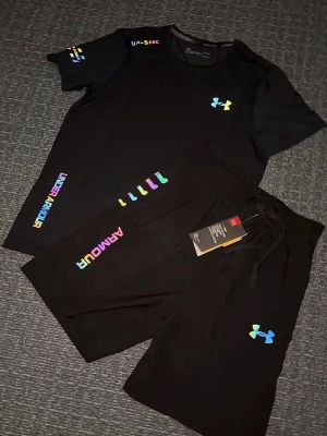 Svart träningsset från Under Armour - Säljer en svart träningsset från Under Armour med färgglada loggor och detaljer. Shortsen har snörning i midjan och är tillverkade i ett mjukt och lätt material som passar perfekt för träning eller chill. Snygg och sportig look med regnbågsfärgade tryck på benet. Storlek S, helt ny 