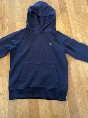 Mörkblå hoodie från Gant - Mörkblå hoodie från Gant med klassisk huva och magficka. Diskret Gant-logga broderad på bröstet. Tillverkad i mjukt bomullsmaterial och har ribbade muddar vid ärmslut och nederkant. Hoodien har små riv sår vid armbågarna men är bara synliga vid när syn.  Strl 146-152