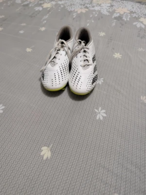 Vita Adidas Predator fotbollsskor - Säljer ett par vita Adidas Predator fotbollsskor med svarta detaljer och snörning. Skorna har ventilerande hålmönster på ovandelen och gula inslag på sulan. Perfekta för dig som vill ha stilrena och funktionella skor på planen.