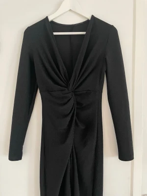 Svart långklänning - Skimrig svart klänning med lång ärm och v-ringning framtill. Klänningen har en snygg twist-detalj i midjan och är figurnära, men stretchig, med en lätt drapering. 