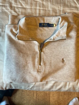 Beige halvzip från Polo Ralph Lauren - Snygg ljusbeige tröja med halv dragkedja från Polo Ralph Lauren. Klassisk broderad logga på bröstet och ribbade muddar vid ärmslut och nederkant. Perfekt till jeans eller chinos för en clean look.