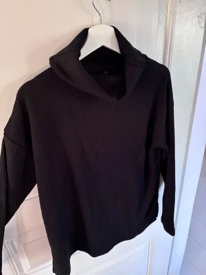 Svart ribbstickad polotröja - Säljer en svart ribbstickad polotröja från Jyothi. Tröjan har en hög, bred krage och långa ärmar. Materialet är mjukt och stretchigt, perfekt för kyligare dagar. Enkel och stilren design som passar till det mesta.