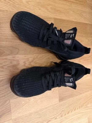 Svarta Nike Air VaporMax sneakers - Säljer ett par svarta Nike Air VaporMax sneakers med stickad ovandel och klassisk snörning. Skorna har en sportig look med synlig Air-sula och diskret Nike-logga på plösen. Perfekta för dig som gillar stilrena och bekväma sneakers.