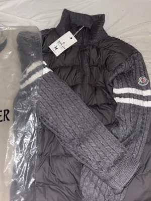 Grå Moncler cardiganjacka med dragkedja - Snygg grå cardiganjacka från Moncler med stickade ärmar och två vita ränder på överarmarna. Kroppen är quiltad och vadderad för extra värme, och jackan har hög krage samt dragkedja framtill. Moncler-logga på vänster ärm. Perfekt för kyliga dagar. Storlek M Helt Ny, säljer för den passa inte mig.