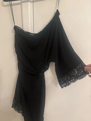 Ny Zara klänning - Svart asymmetrisk klänning med oneshoulder och tunna axelband. Klänningen har spetsdetaljer längs kanterna och en snygg cut-out vid midjan. Materialet är mjukt och följsamt, perfekt för en trendig look.