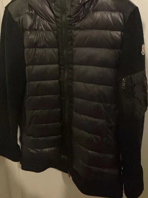 Svart dunjacka med huva - Snygg svart dunjacka med quiltad framsida och huva. Jackan har dragkedja framtill, släta ärmar och en ficka med dragkedja på ena ärmen. Perfekt för kalla vinterdagar och har en modern, sportig look.