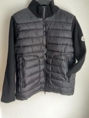 Svart dunväst med stickade ärmar - Snygg svart dunväst med quiltad framsida och två dragkedjeförsedda fickor. Västen har ribbstickade ärmar och hög krage. Moncler-logga på ena ärmen. Perfekt för lager-på-lager och streetstyle.
