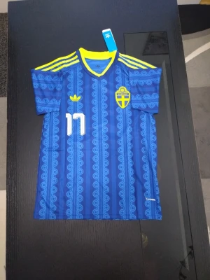 Blå Sverige fotbollströja Adidas - Säljer en blå fotbollströja från Adidas med Sveriges landslagsemblem och nummer 11 på bröstet. Tröjan har gula detaljer, klassiska Adidas-ränder på axlarna och ett retroinspirerat mönster. Perfekt för dig som vill stötta Sverige med stil.