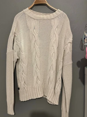Beige kabelstickad tröja - Snygg beige stickad tröja med grova kabelstickade detaljer framtill. Tröjan har rund halsringning och långa ärmar. Perfekt för en ”downtown girl” stil. Lite mer åt det oversize hållet, 100% bomull.