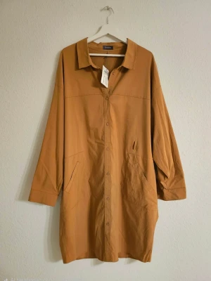 Skjortklänning Samoon - Stilren beige skjortklänning med knappar framtill och två stora fickor. Klänningen har lång ärm med knappdetaljer vid ärmslut och klassisk krage. Perfekt för en avslappnad men trendig look. Nypris: 1699 KR