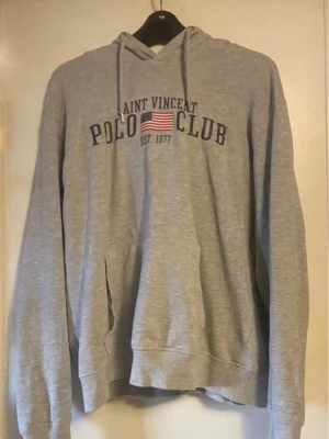 Grå hoodie Saint Vincent Polo Club - Grå hoodie med tryck från Saint Vincent Polo Club och amerikansk flagga på bröstet. Klassisk modell med huva och dragsnören samt magficka framtill. Tillverkad i mjukt sweatshirtmaterial, perfekt för chill dagar.lite trasig under armen 