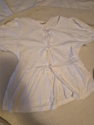 Vit blus med knyt och kort ärm - Söt vit blus med tre knyt framtill och korta puffärmar. Blusen har en lätt utsvängd nederdel och mjukt fallande material. Perfekt för en sommardag 