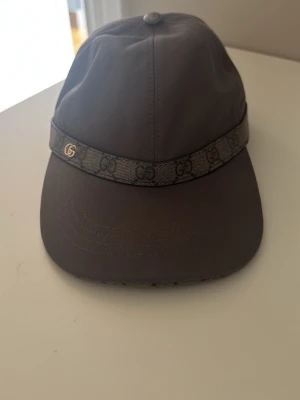 Mörkbrun keps från Gucci - Snygg mörkbrun keps från Gucci med klassiskt GG-mönsterband runtom och en liten guldfärgad GG-detalj på sidan. Kepsen har böjd skärm och justerbar rem bak. Materialet ser ut att vara en blandning av textil och syntet.