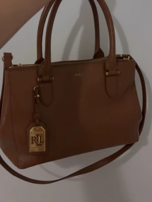 VINTAGE Brun handväska från Ralph Lauren - Original pris: 5,999- 7000 kr Snygg brun handväska i skinn från Lauren Ralph Lauren med guldfärgade detaljer och RL-logga. Väskan har både handtag och axelrem, perfekt för att bära på olika sätt. Klassisk och stilren design med dragkedja upptill.