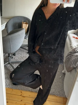 Svart loungewear-set med skjorta - Säljer ett svart loungewear-set med avslappnad skjorta och matchande byxor. Skjortan har knappar framtill och en lös passform med långa ärmar. Byxorna är vida och har resår i midjan för extra komfort. Perfekt för chill dagar hemma.