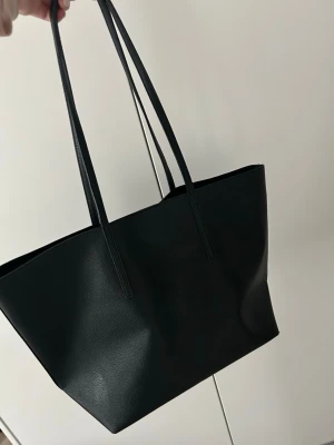 Svart shoppingväska i konstläder Mango - Stilren svart shoppingväska från Mango med långsmala axelband och rymlig insida. Väskan är tillverkad i slätt konstläder och har en enkel, modern design utan synliga loggor eller detaljer. Perfekt för dig som gillar minimalistisk stil och behöver plats för allt du vill bära med dig.