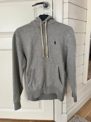 Grå hoodie från Polo Ralph Lauren XS - Snygg grå hoodie från Polo Ralph Lauren med klassisk huva och vita dragsnören. Har en känguruficka framtill och den ikoniska lilla mörkblå loggan broderad på bröstet. Tillverkad i mjukt sweatshirtmaterial, perfekt för en chill och avslappnad stil. Storlek Xs