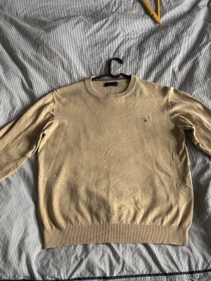 Beige stickad tröja från Gant - Snygg beige stickad tröja från Gant med rund halsringning och diskret broderad logga på bröstet. Tröjan har ribbade muddar vid ärmslut och nederkant. Perfekt för lager på lager och enkel att matcha med olika outfits.            Passar cirka 170 cm. 
