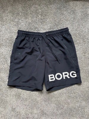 Svarta badbyxor från BORG - Snygga svarta badbyxor från BORG med vit logga på vänster ben. Klassisk modell med resår och dragsko i midjan samt sidofickor. Tillverkade i snabbtorkande material, perfekta för stranden eller poolen.