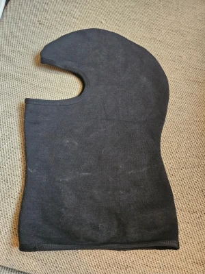 Svart balaklava i bomull - En svart balaklava i mjukt bomullstyg. Klassisk modell med öppning för ansiktet och täcker både huvud och hals. Perfekt för kalla dagar eller som extra lager under hjälm. Enkel och stilren design utan synligt märke.