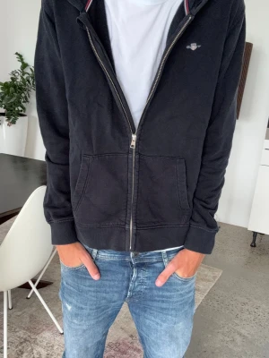 Svart hoodie med dragkedja från gant - Svart hoodie med dragkedja framtill och två stora fickor. Klassisk huva och ribbade muddar vid ärmslut och nederkant. Liten diskret logga på bröstet och detaljer i rött och vitt vid huvan. Perfekt till jeans. Storlek 176