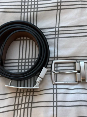 Ralph lauren bälte svart/brun - Stilrent svart bälte från Polo med två olika silverfärgade spännen, ett klassiskt och ett med Polo-loggan ingraverad. Bältet är i slätt skinn och passar perfekt till jeans eller kostymbyxor. Det går att vrida om spännet så att man kan använda både den bruna stilen eller den svarta. Skärpet har två extra hål se sista bilderna. Hör av dig vid frågor 
