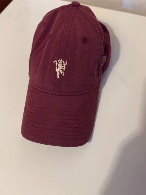 Vinröd Manchester United keps - Snygg vinröd keps från New Era med Manchester United-logga broderad framtill. Klassisk 9FORTY-modell med böjd skärm och justerbar passform. Tillverkad i bomullstyg och har klubbmärke på insidan.