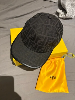 Svart keps med Fendi-logga - Snygg svart keps från Fendi med det klassiska FF-monogrammet över hela kepsen. Kepsen har böjd skärm med läderdetalj och justerbar rem bak. Tillverkad i tyg och levereras med gul Fendi-påse och box.