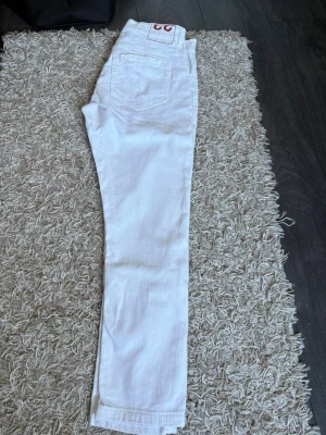 Vita skinny jeans med bakficka - Säljer ett par vita skinny jeans med klassisk femficksdesign och en liten patch med röd logga bak. Jeansen har smal passform och raka ben, perfekta till en clean och fräsch look. Materialet är mjukt och stretchigt för extra komfort. Tyvärr finns inget kvitto, dom är 93cm i längden och 38cm i midjan.
