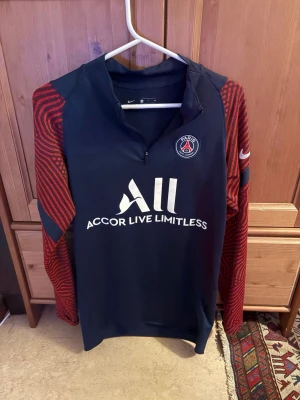 PSG träningströja från Nike - Mörkblå PSG träningströja från Nike med röda mönstrade ärmar och halv dragkedja. Paris Saint-Germain-logga på bröstet och vit ACCOR LIVE LIMITLESS-tryck framtill. Tillverkad i ett lätt och funktionellt material, perfekt för träning.