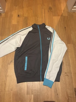 Svart och vit track jacket Fred Perry - Snygg vintage track jacket från Fred Perry i svart med vita ärmar och blå detaljer. Jackan har dragkedja framtill, ribbade muddar och blå logga på bröstet. Perfekt för en sportig och retro look. Materialet är 50% bomull och 50% polyester.