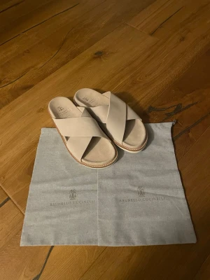 Beige sandaler Brunello Cucinelli - Stilrena beige sandaler från Brunello Cucinelli med breda korslagda remmar i mjukt läder. Sandalerna har en klassisk slip-in design och en ljus sula med kontrastsömmar. Perfekta för en avslappnad och trendig look under varma dagar.