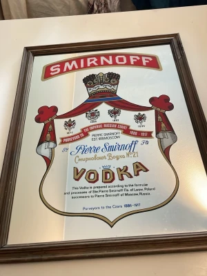 Smirnoff Vodka spegel med ram - Snygg spegel med Smirnoff Vodka-motiv i färgerna röd, guld, blå och vit. Spegeln har en brun träram och tryckt text samt illustrationer av krona och banderoller. Perfekt som dekoration till baren eller köket spegeln e då ifrån 1970-1980 talet i grymt bra skick.