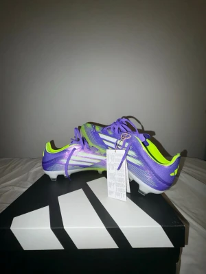 Lila och gula Adidas fotbollsskor - Snygga fotbollsskor från Adidas i lila och gult med vita detaljer. Skorna har klassisk snörning, låg profil och är tillverkade i ett lätt syntetmaterial för bästa känsla på planen. Perfekta för dig som vill sticka ut på matchen.