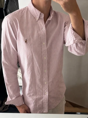 Rosa och vit randig skjorta - Säljer en klassisk rosa/röd och vit randig skjorta med button-down krage och långa ärmar. Skjortan har knappar framtill och en liten broderad logga på bröstet. Perfekt för en stilren och avslappnad look.