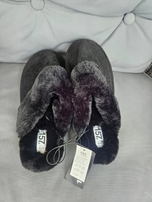 Svarta fluffiga tofflor med fuskpäls - Mysiga svarta tofflor från 157 med mjuk fuskpäls runt kanten och ovansida i mockaimitation. Perfekta för att hålla fötterna varma hemma. Klassisk slip-in modell med rund tå och platt sula. Storlek 40/41  Kan även hämtas I Partille