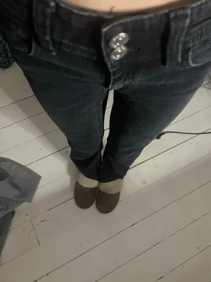 Svarta utsvängda jeans med dubbla knappar - Säljer ett par svarta jeans med raka ben och dubbla silverknappar i midjan. Jeansen har klassisk femficksmodell och är tillverkade i ett stretchigt denimtyg som sitter snyggt. Perfekta för dig som gillar en enkel men stilren look.