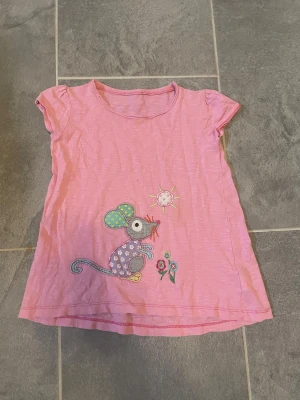 Rosa t-shirt med musapplikation för barn - Söt rosa t-shirt med korta ärmar och en färgglad applikation av en mus, blommor och sol på framsidan. Tillverkad i mjuk bomull som känns skön mot huden. Perfekt för dig som gillar gulliga och lekfulla motiv.