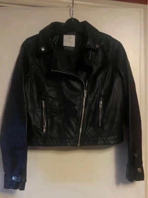 Svart skinnjacka med dragkedjor(barn)  - Snygg svart skinnjacka i bikerstil med sned dragkedja framtill och två dragkedjeförsedda fickor. Jackan har klassisk krage med tryckknappar och detaljerade sömmar på ärmarna. Perfekt för en cool och trendig look.