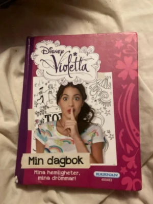 Disney Violetta Min dagbok - En färgglad dagbok med hårda pärmar från Disney Violetta. Omslaget är lila och pryds av Violetta och olika mönster. Perfekt för att skriva ner hemligheter, drömmar och tankar. Boken har illustrationer och foton på karaktärer från serien.lite sliten på utsidan 
