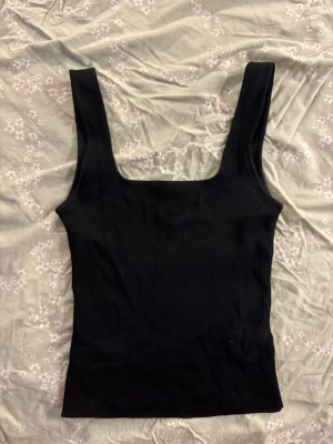 Svart ribbad linnetopp - Snygg svart ribbad linnetopp med breda axelband och fyrkantig ringning. Toppen är tight och har en enkel, stilren design som passar perfekt till jeans eller kjol. Perfekt för dig som gillar en basic men trendig look.