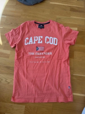 Korallröd CAPE COD t-shirt medium - Snygg korallröd t-shirt med trycket 'CAPE COD' i vitt framtill, samt en liten flagga och textdetaljer. T-shirten har rund hals och korta ärmar. Perfekt för en avslappnad och cool stil. Materialet känns mjukt och skönt mot huden. Medium