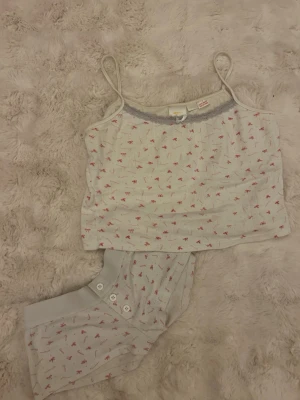 Söt pyjamas från Gina Tricot - Pyjamas-set med linne och byxa från Gina Tricot. Vit bas och små rosa rosetter och spetskant upptill. Linnet har smala axelband och en liten rosett framtill. Byxan har knappar vid benslutet. Perfekt för mysiga kvällar.