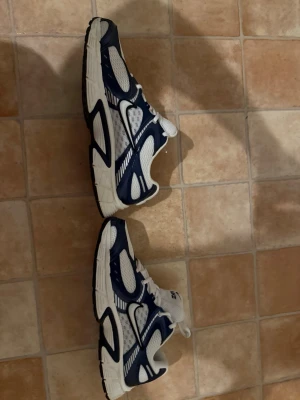Nike vita och marinblå sneakers - Säljer ett par snygga Nike sneakers i vitt och marinblått. Skorna har en sportig design med meshpaneler för bra ventilation och klassiska snörningar. Perfekta för dig som gillar retrostil och vill ha bekväma skor till vardags. Nike-logga på plösen och sidan.