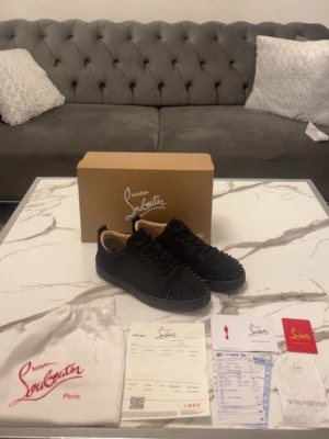 Svarta Louboutin sneakers med nitar - Säljer ett par svarta sneakers från Christian Louboutin i mocka med coola svarta nitar över hela tån och sidorna. Skorna har en låg silhuett, svart sula och klassisk röd detalj på insidan. Kommer med originalkartong, dustbag och äkthetsbevis.